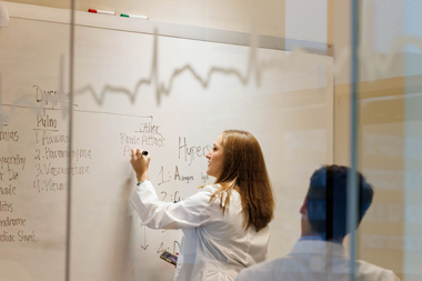 In study room, one resident writes on the whiteboard while another looks on while seated.